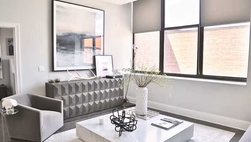Bright modern living room with large windows, a gray armchair, geometric sideboard and a marble coffee table with decorative vase.