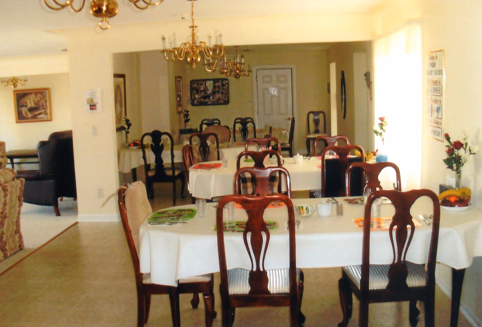Dining room with multiple tables covered with white tablecloths and set with placemats, utensils, and condiments. Wooden chairs surround the tables. The room has chandeliers hanging from the ceiling and a window with sheer curtains letting in natural light. There are flowers and fruit bowls on the tables and walls decorated with pictures and signs.