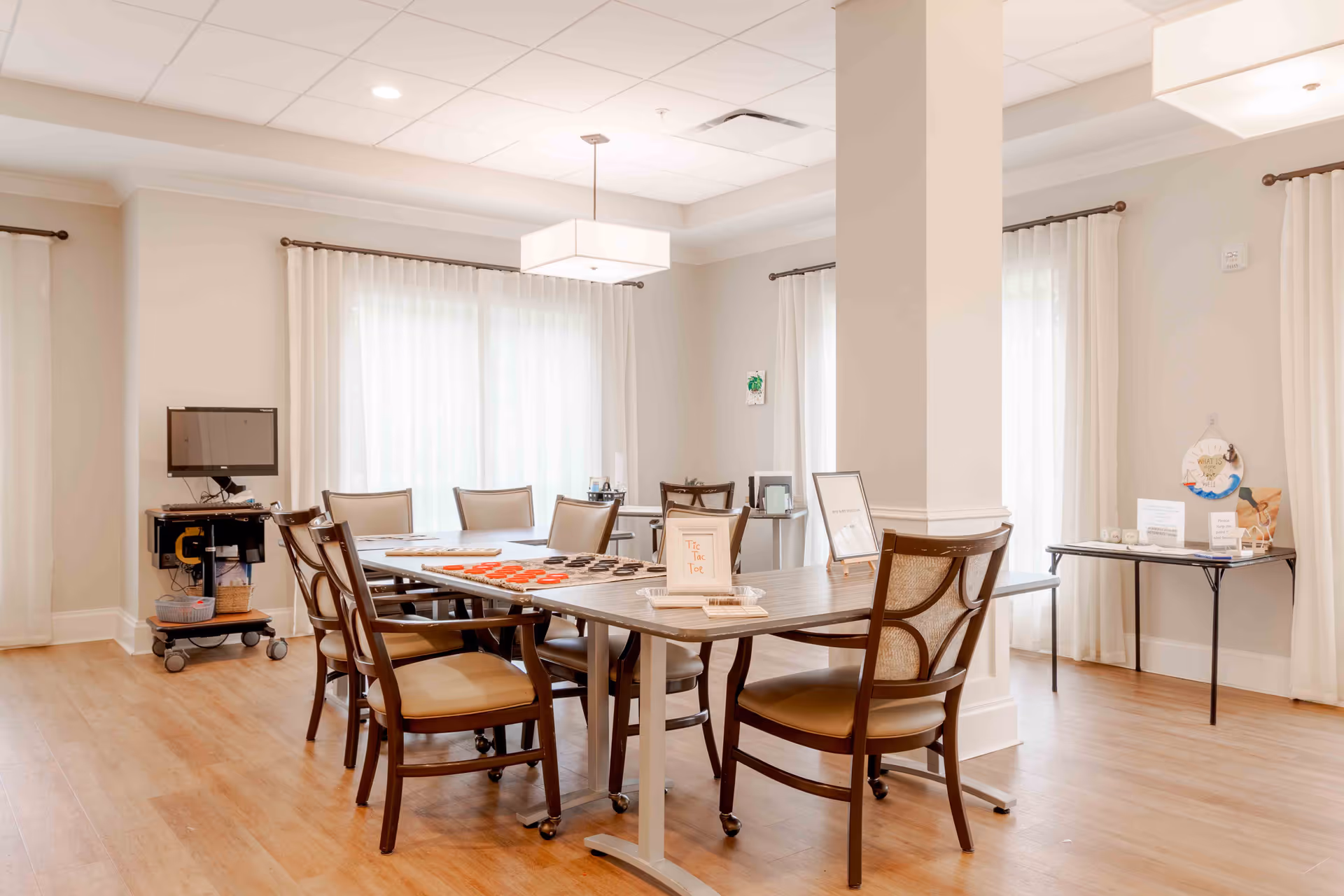 Bright communal activity/dining room with a large table surrounded by chairs, game pieces on the table, and a TV cart.
