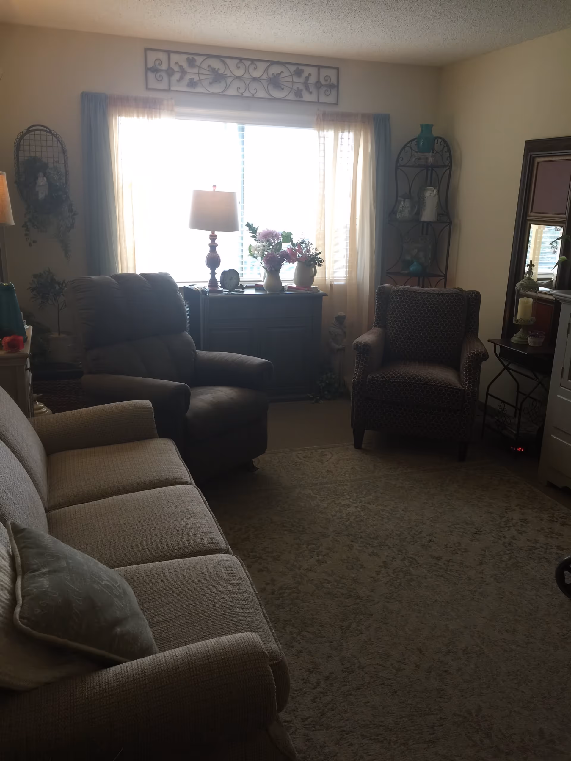 A cozy living room with a beige sofa, a dark recliner chair, and a patterned armchair. A window with sheer curtains lets in natural light behind a small cabinet topped with a table lamp and flower vases. Decorative items and plants are placed around the room, including a metal shelf with vases and a large mirror on the wall.