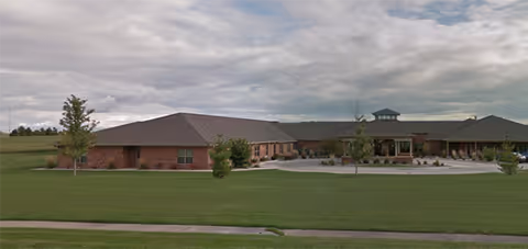 Wide exterior view of a single-story brick building with a large, well-maintained green lawn in front under a cloudy sky. The building has a low-pitched roof and a covered entrance area with columns.