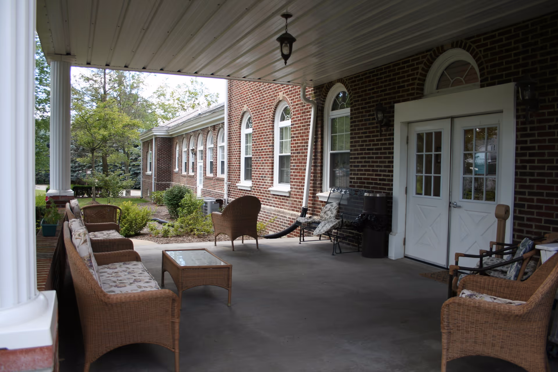 Covered outdoor patio area with wicker furniture including a sofa, chairs, and a glass-top coffee table. The patio is attached to a brick building with white-framed windows and double doors. There are some plants and trees visible in the background.