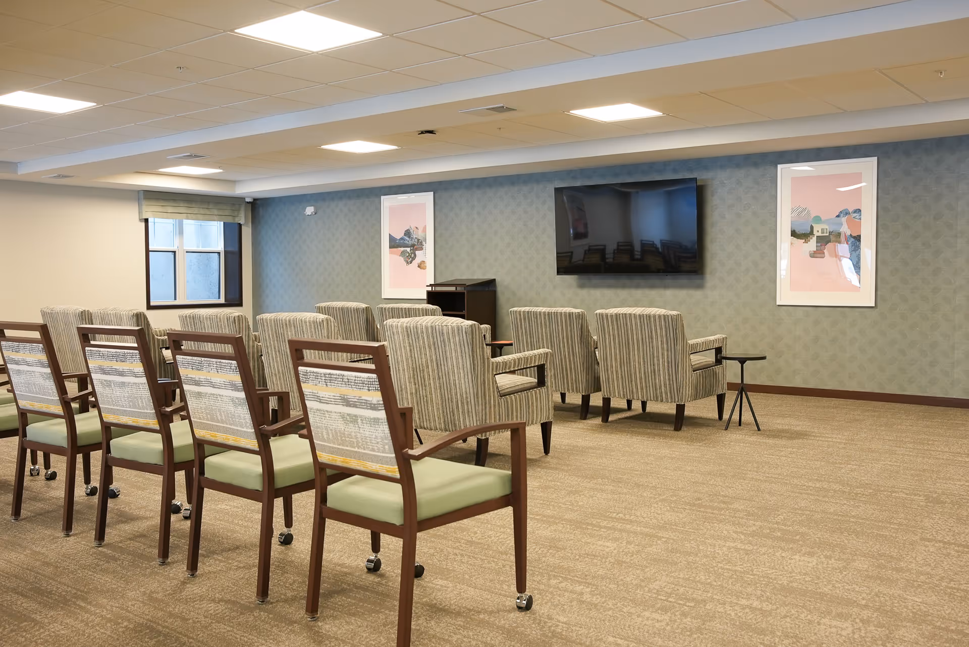 A senior living facility common area with rows of chairs facing a large wall-mounted flat screen TV. The room has a carpeted floor, patterned wallpaper, two framed artworks on the wall, and a window with a shade partially drawn.