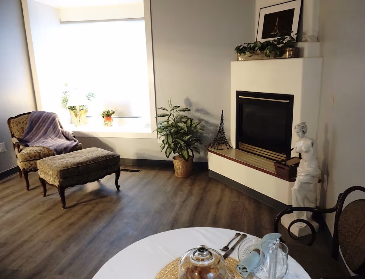 A cozy living room area with a vintage armchair and matching ottoman near a large window with potted plants on the windowsill. There is a fireplace with decorative items including a small Eiffel Tower model and a white statue of a woman. A round table with a white tablecloth, glassware, and utensils is partially visible in the foreground.