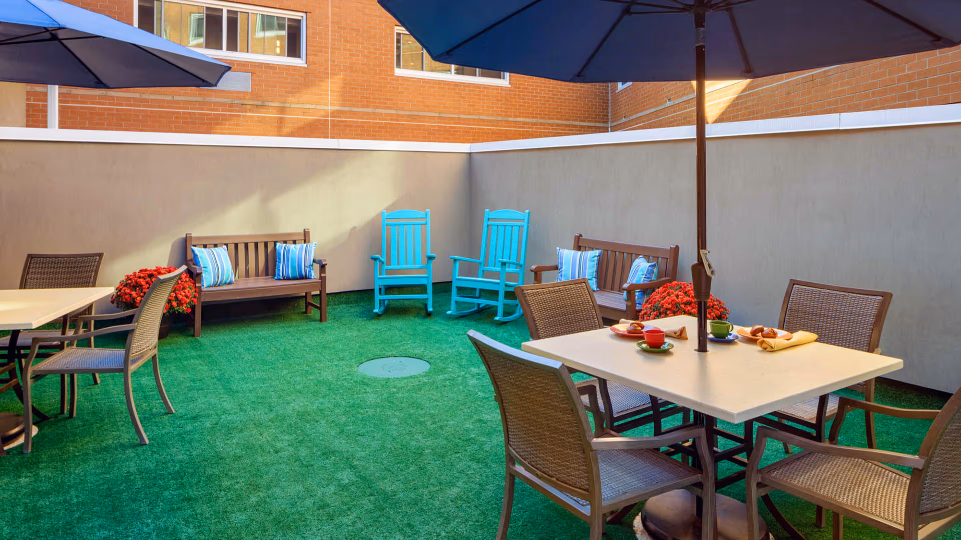 Outdoor patio area with artificial green grass, two wooden benches with blue and white striped pillows, two turquoise rocking chairs, and a table with four chairs under a large blue umbrella. There are red flower pots placed near the benches and the area is enclosed by beige walls with a brick building visible in the background.