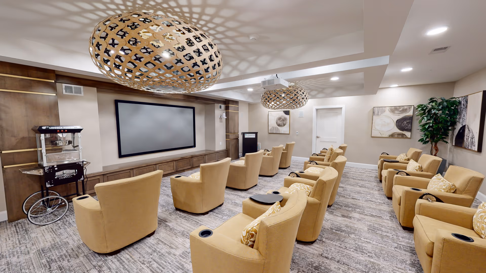 A senior living facility media room with multiple beige recliner chairs arranged in rows facing a large flat screen TV mounted on a wooden accent wall. The room features two large decorative pendant lights hanging from the ceiling, a popcorn machine on the left side, framed artwork on the walls, and a potted plant in the corner. The carpet has a modern pattern and the room is well-lit with recessed lighting.
