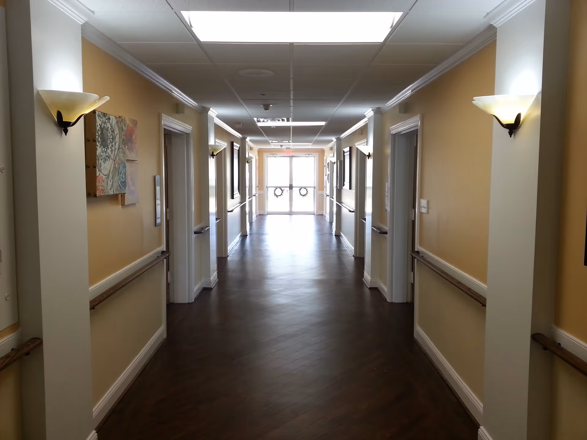 A well-lit hallway in a retirement community with wooden floors, beige walls, handrails on both sides, wall-mounted light fixtures, and several doors leading to rooms. At the end of the hallway, there is a glass door with wreath decorations.