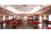 A spacious dining room with multiple tables covered in red tablecloths and chairs arranged around them. The room has a wooden floor, ceiling fans, and is decorated with garlands hanging along the ceiling edges. Large windows with curtains allow natural light to enter the room.