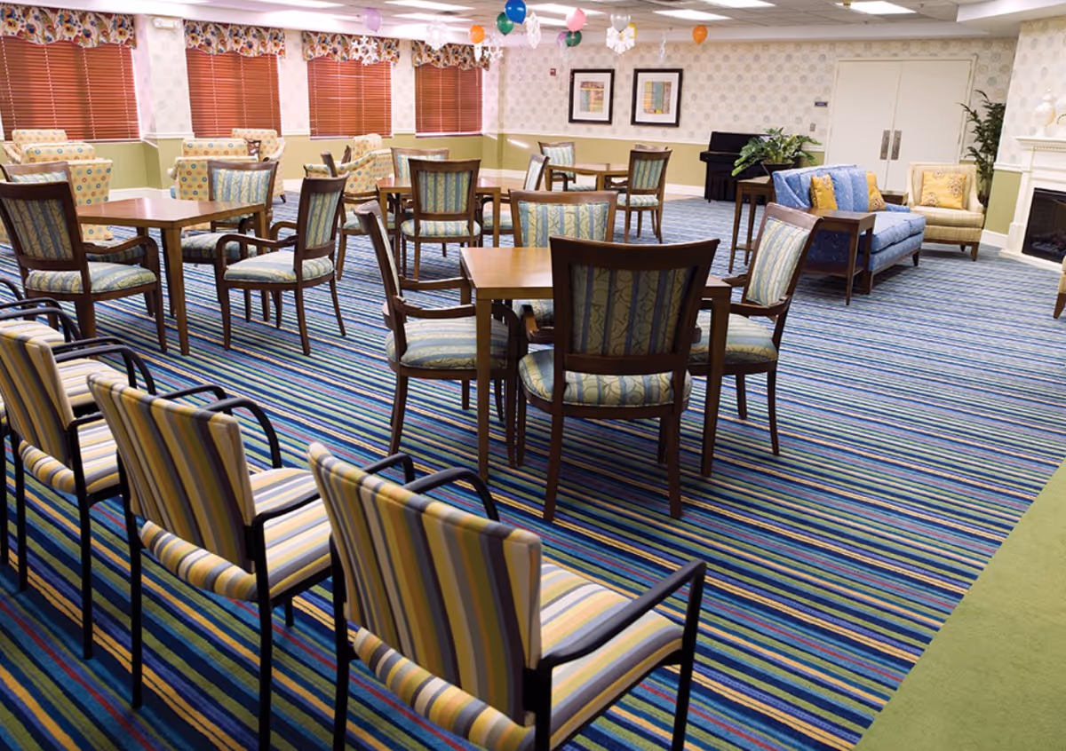 Bright communal dining and activity room with multiple wooden tables, patterned chairs, sofas and a striped carpet.