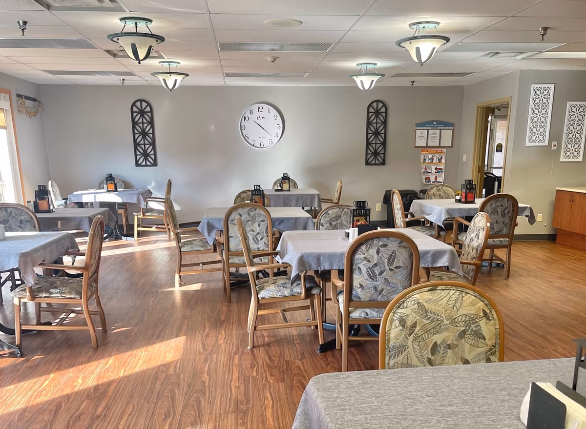 Dining room with multiple tables covered in gray tablecloths and patterned chairs, wood-look flooring and a wall clock.