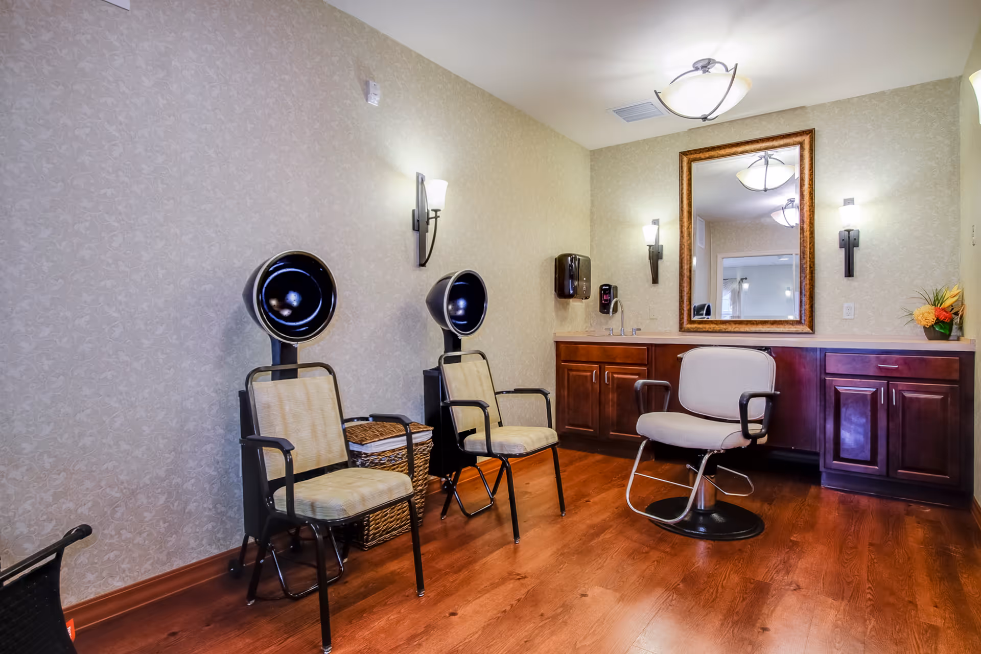 Interior view of a salon area in an assisted living facility with two vintage hair dryers on chairs, a salon chair in front of a large mirror, wooden cabinets, and soft wall lighting.