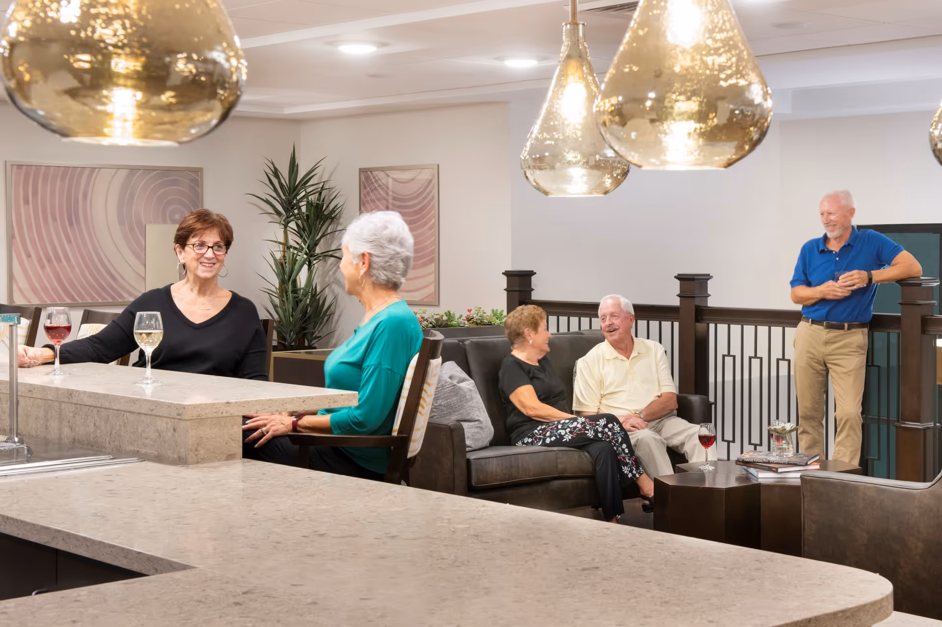 A group of four elderly people socializing in a modern common area with a countertop and comfortable seating. Two women sit at the counter with glasses of wine, while a man and woman sit on a couch laughing. Another man stands nearby smiling. The room is decorated with hanging pendant lights, abstract wall art, and a tall plant.