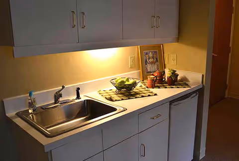 Kitchenette with a stainless steel sink, white cabinets, and a countertop decorated with a fruit bowl and a framed picture.