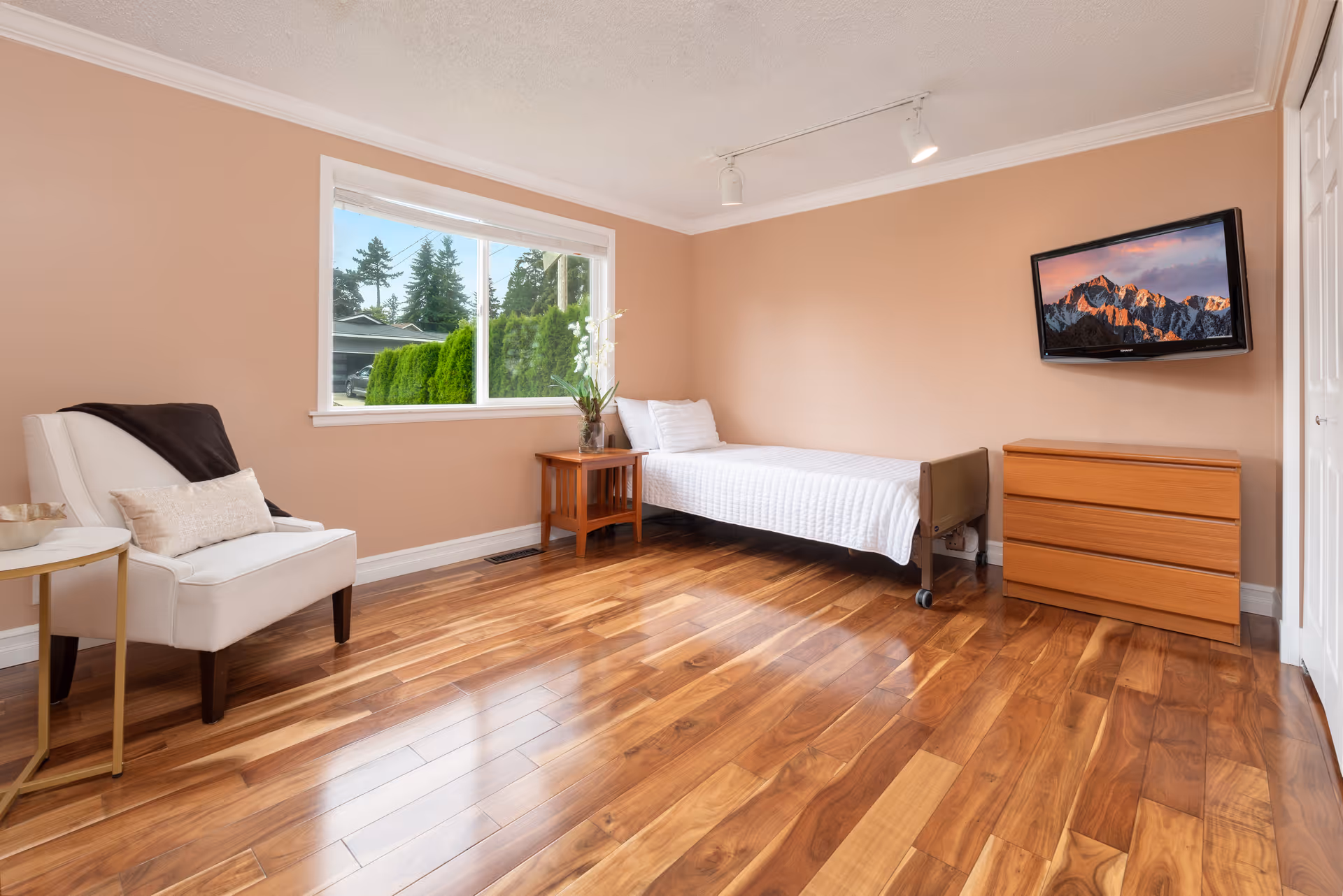 A simple, clean bedroom with a single bed covered in a white quilt, a wooden dresser, a small wooden side table with a vase of flowers, a white armchair with a cushion and a dark throw, and a wall-mounted flat screen TV displaying a mountain landscape. The room has light brown walls, a large window showing greenery outside, and polished wooden floors.