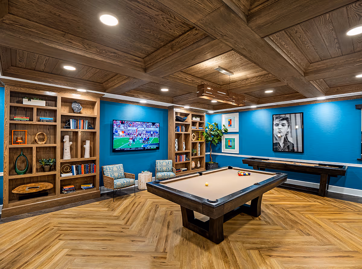 A recreational room with a pool table in the center, a shuffleboard table against a bright blue wall, two patterned chairs with a small wooden stump table between them, built-in wooden bookshelves filled with books and decorative items, a flat-screen TV mounted on the wall showing a football game, and wooden coffered ceiling with recessed lighting.