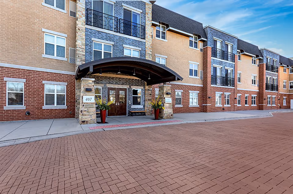 Exterior front entrance of a multi-story senior living facility with a covered entryway supported by stone pillars, large windows, and a brick-paved driveway in front.