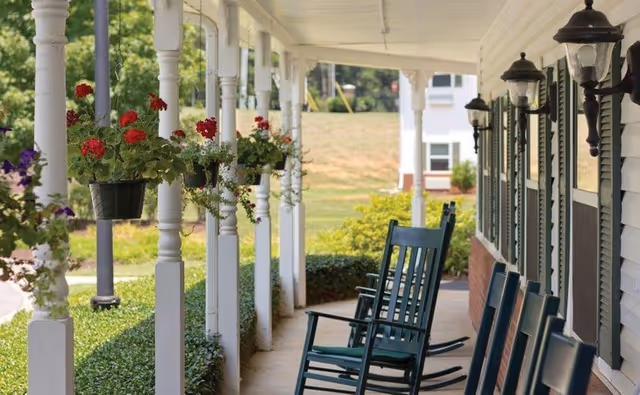 A covered porch with white columns and hanging flower pots filled with red and purple flowers. Several dark green rocking chairs are lined up along the porch next to a white building with black shutters and wall-mounted lantern lights. The background shows a grassy area and another building in the distance.