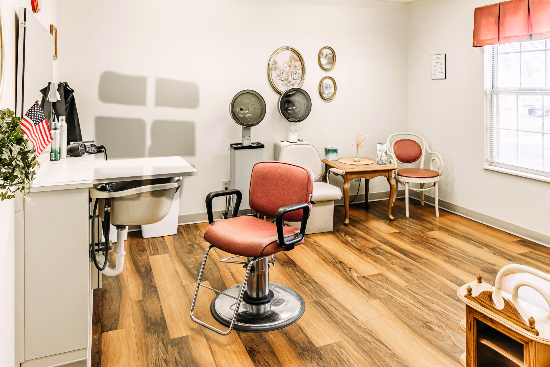 A bright hair salon room with a red salon chair in the center, two vintage hair dryers against the wall, a small wooden table with a vase of flowers, and two chairs with red cushions. The room has wood flooring, a window with blinds and a red valance, and an American flag on the left side near a sink and countertop.