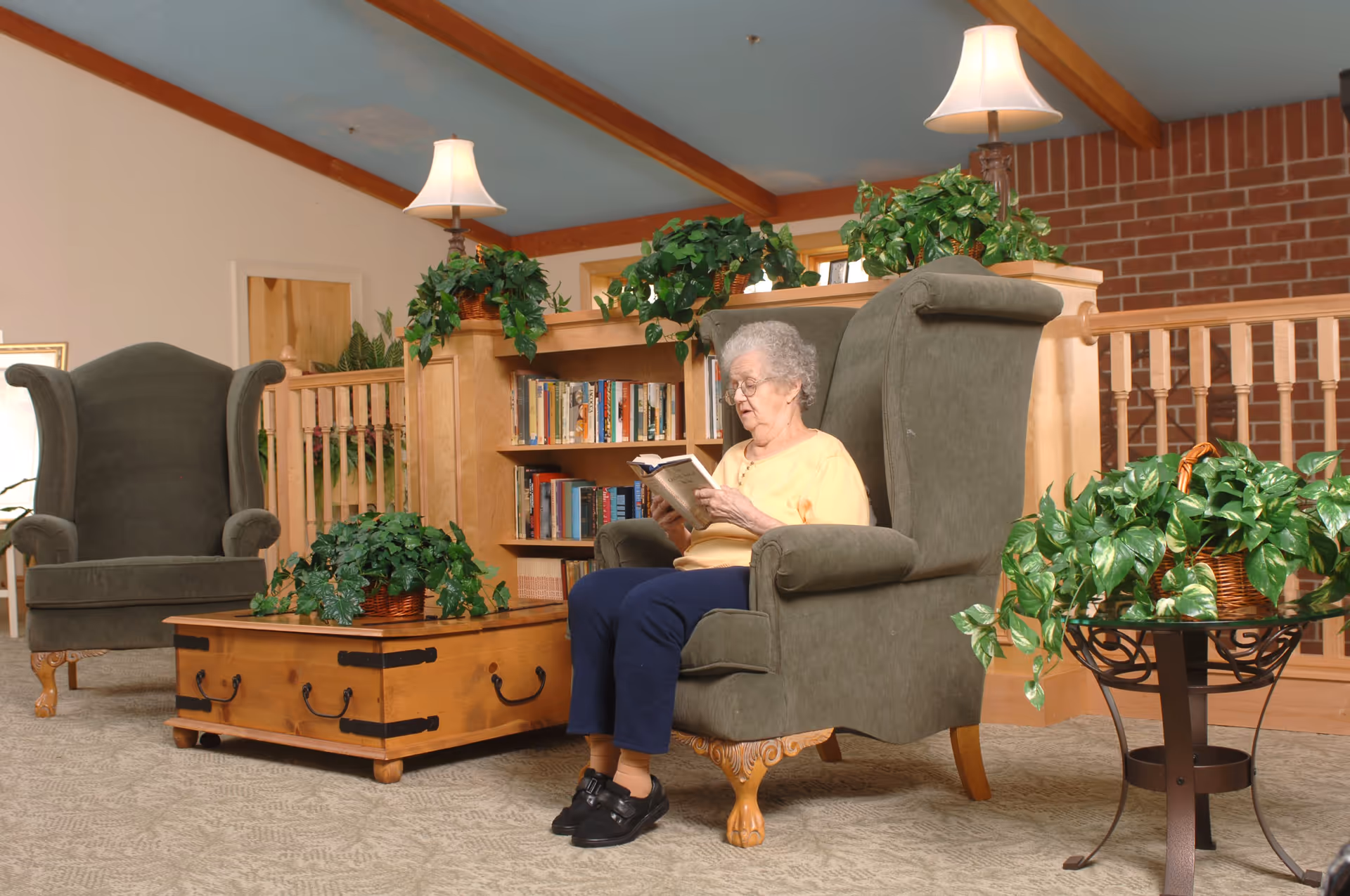 An elderly woman with gray hair and glasses sits in a green upholstered armchair reading a book in a cozy living room. The room features a wooden coffee table with a potted plant, another matching armchair, a bookshelf filled with books, several green potted plants, two lamps with white shades, a brick wall, and wooden ceiling beams.