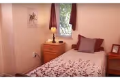 Small furnished bedroom with a single bed, nightstand, lamp, window, and wall art.