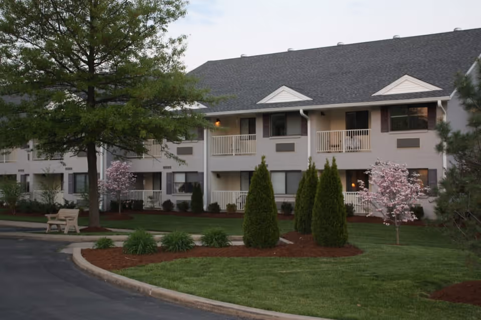Exterior view of a two-story senior living facility building with balconies, surrounded by well-maintained landscaping including trees, bushes, and flowering plants. A paved driveway and a bench are visible in the foreground.