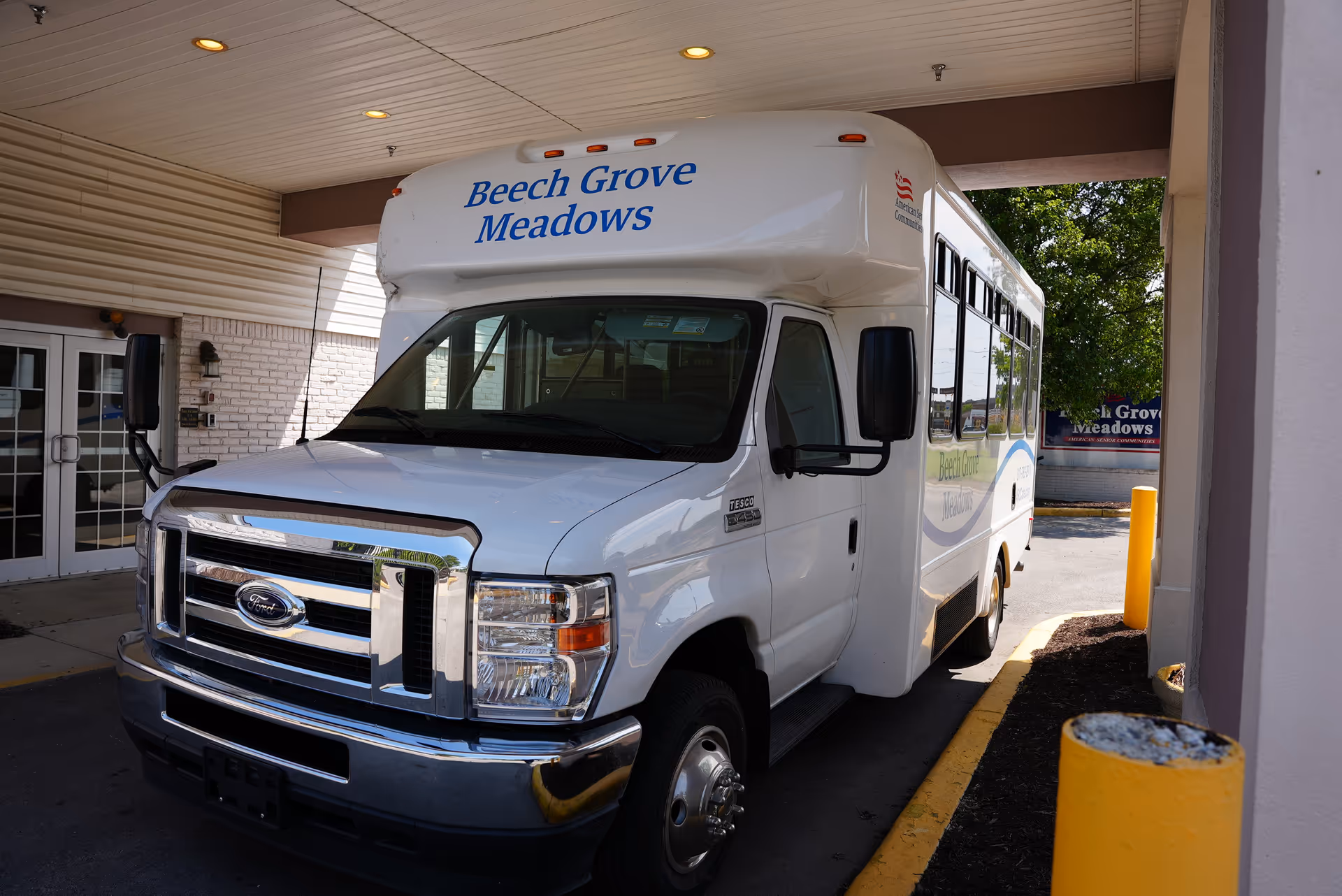 A white shuttle bus parked under a covered entrance with the name 'Beech Grove Meadows' displayed on the front and side of the bus. The bus is a Ford model and is positioned near a building entrance with glass doors and yellow bollards nearby.