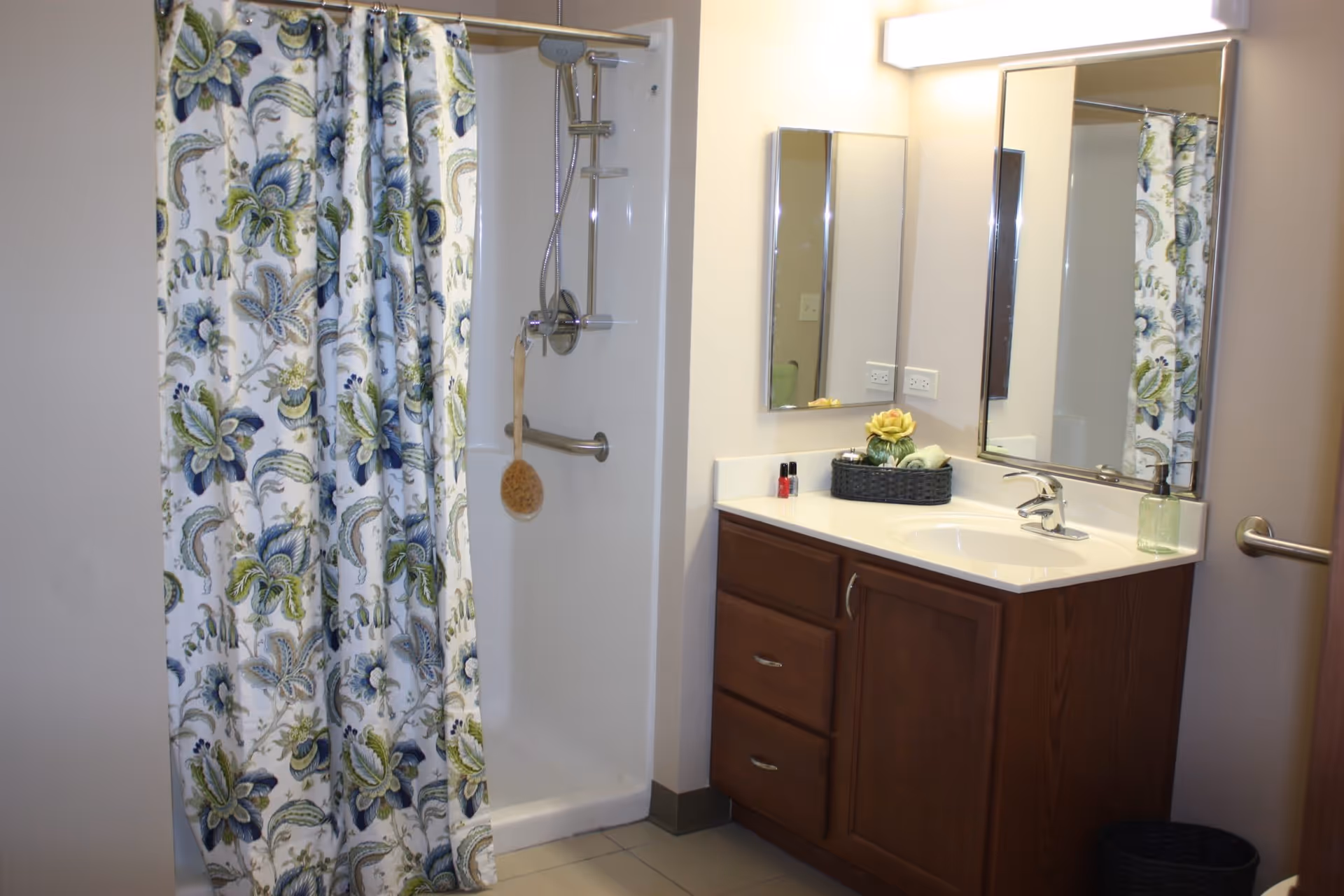 Bathroom with a floral patterned shower curtain, a walk-in shower with a handheld showerhead and grab bar, a wooden vanity with a white countertop, a sink, two mirrors, and a basket with toiletries on the counter.