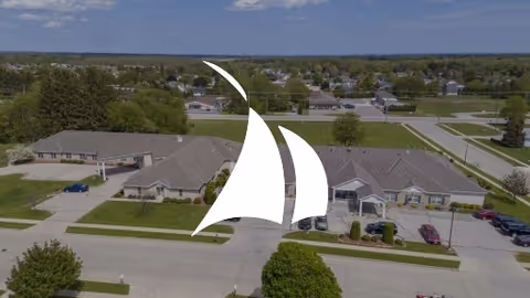 Aerial exterior view of a single-story assisted living complex and nearby streets, overlaid with a white sail-shaped logo.