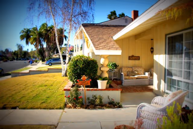 Front porch of a single-story house with a small garden area, a wicker sofa with cushions, a white wicker chair, and a decorative sign hanging on the wall. The house has a sloped roof and is painted yellow. The street and neighboring houses are visible in the background under a clear blue sky.