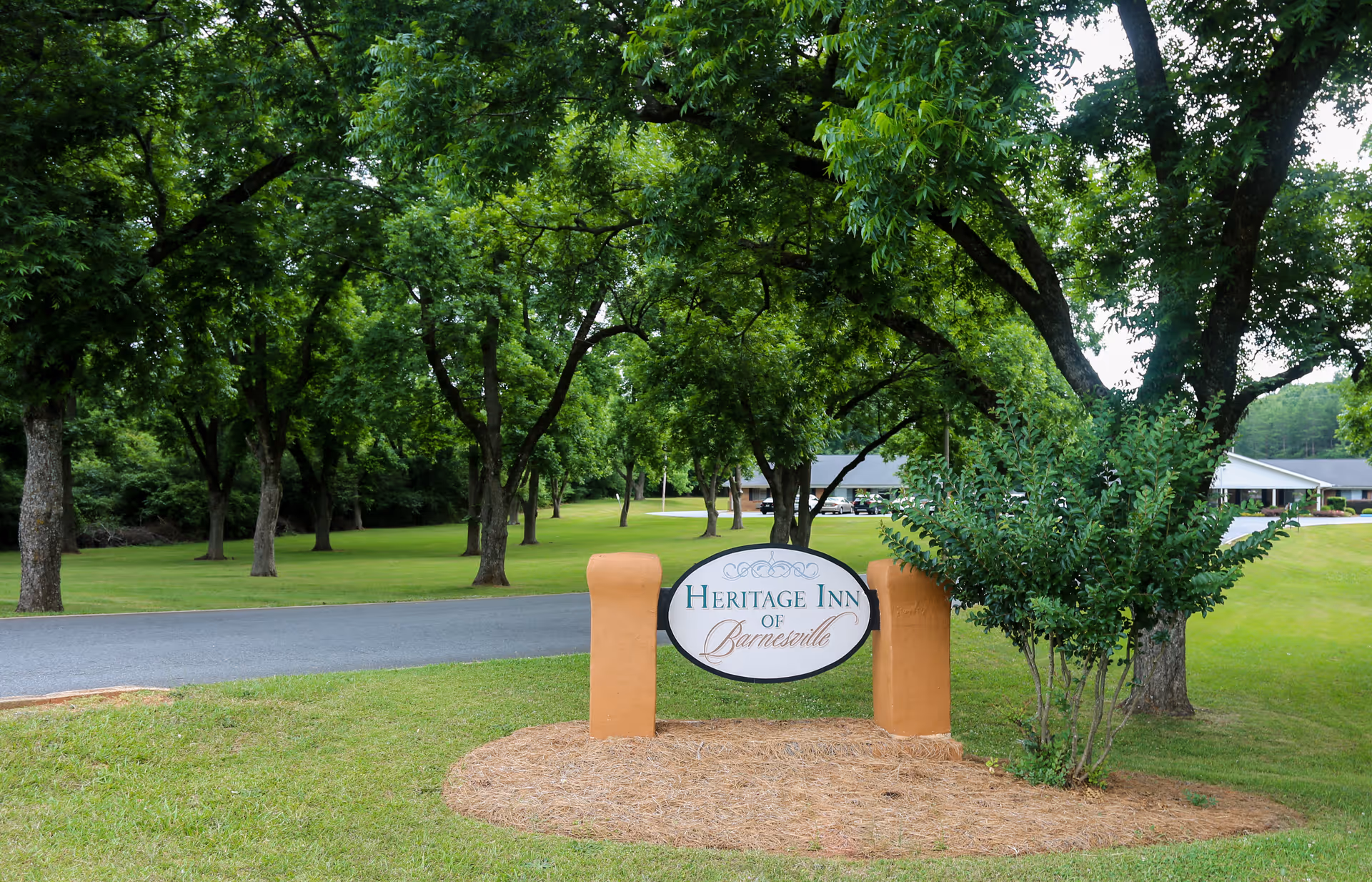 Outdoor view of the entrance area of Heritage Inn of Barnesville, featuring a sign with the facility's name surrounded by green grass, trees, and a paved driveway leading to buildings in the background.