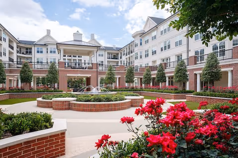Courtyard with a central fountain and brick planters surrounded by a multi-story senior living building, with red flowers in the foreground.