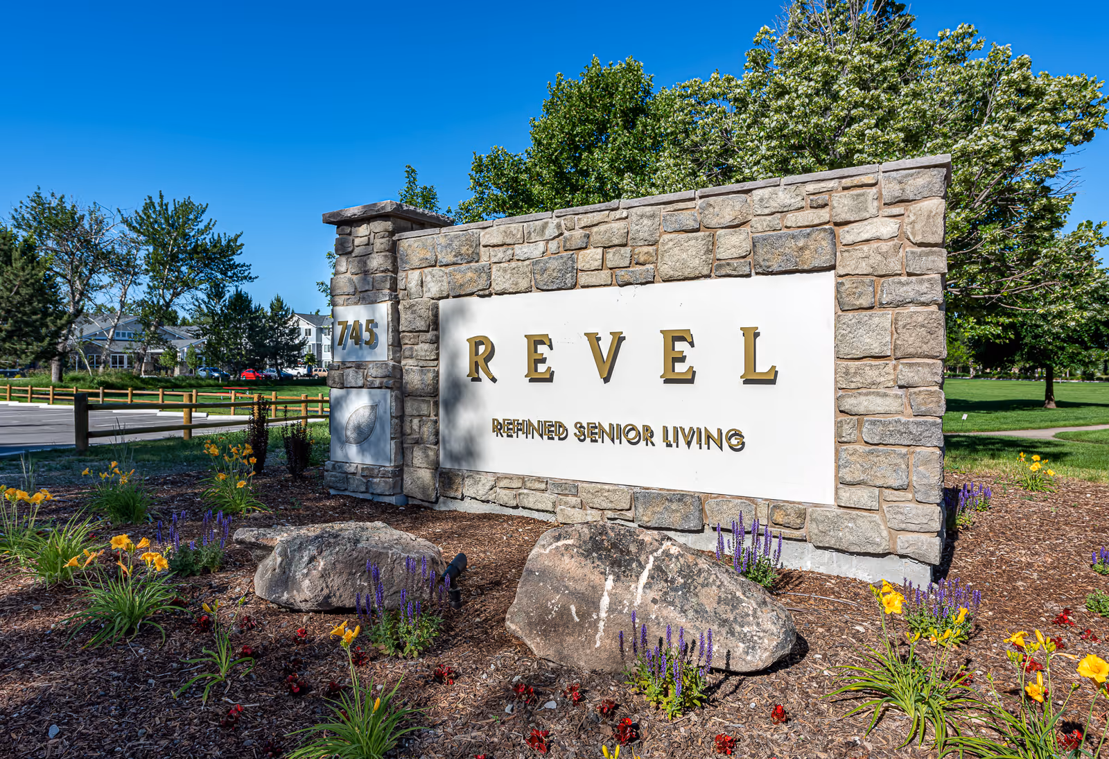 Stone entrance sign reads REVEL with Refined Senior Living lettering, surrounded by flowers.