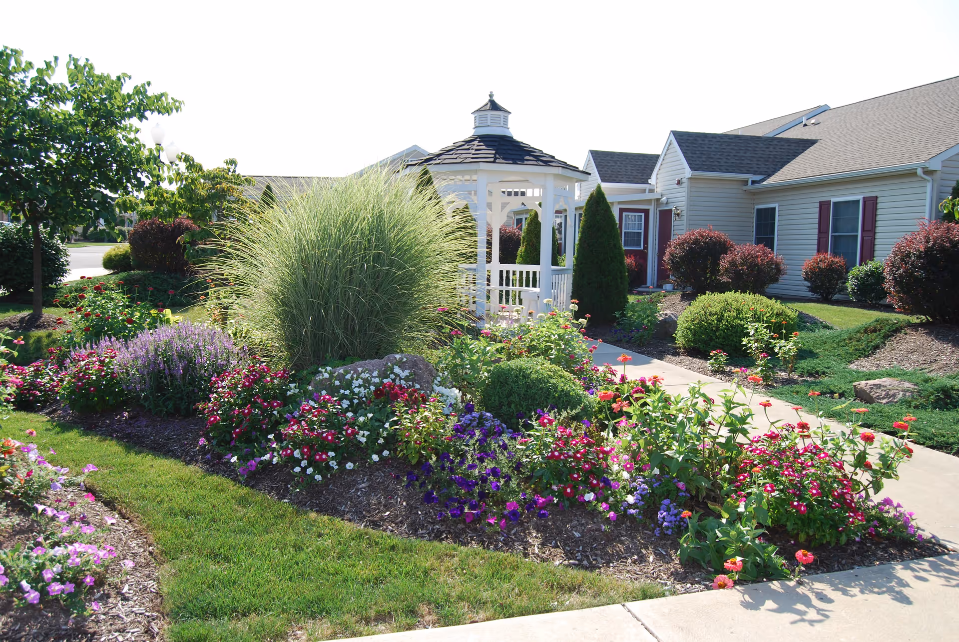 Landscaped garden with colorful flower beds and a white gazebo in front of a single-story senior living building.