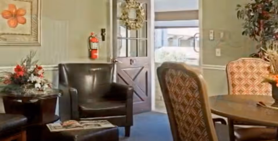 A cozy interior room with a dark leather armchair and ottoman, a round wooden table with patterned upholstered chairs, floral arrangements on the table and side table, a door with glass panes decorated with a wreath, and a fire extinguisher mounted on the wall.