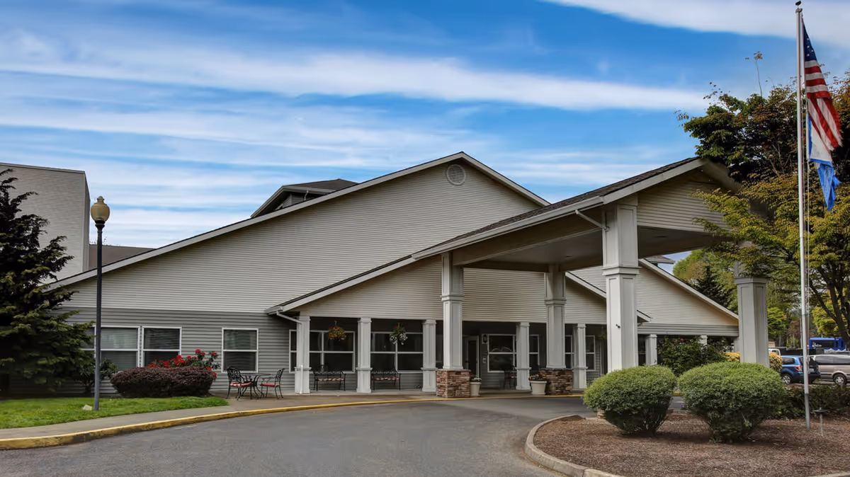 Entrance facade of a single-story senior living building with a covered drive-up portico, landscaping, and an American flag.