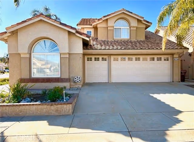 Front exterior of a beige stucco house with a three-car garage, tiled roof, driveway, and small landscaped planter with palm trees.