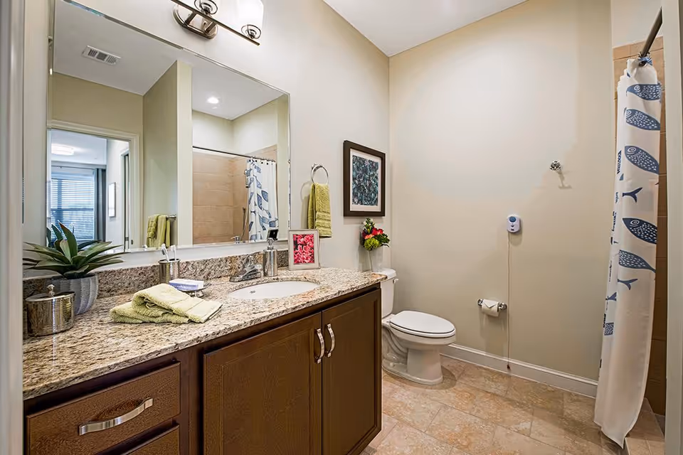 A clean and modern bathroom featuring a granite countertop with a sink, a large mirror above the sink, a toilet, and a shower with a curtain decorated with blue fish patterns. The bathroom has beige walls and tiled flooring. There are green towels, a small plant, framed pictures, and a soap dispenser on the countertop.