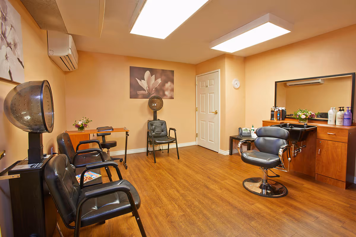 Interior of a hair salon room with wooden flooring and beige walls. The room contains black salon chairs, a hair dryer, a sink with hair care products on a wooden cabinet, a small table with a telephone, and floral wall art. The ceiling has large rectangular fluorescent lights.
