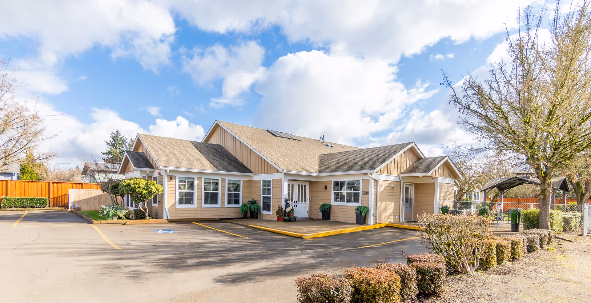 Exterior view of a single-story residential building with beige siding and multiple windows, surrounded by a parking lot with marked spaces and landscaped bushes and trees under a partly cloudy sky.