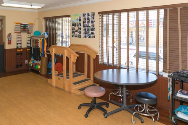 A rehabilitation room with wooden stairs for physical therapy, exercise equipment including weights and balls, a round table with two stools, and large windows with wooden blinds letting in natural light.