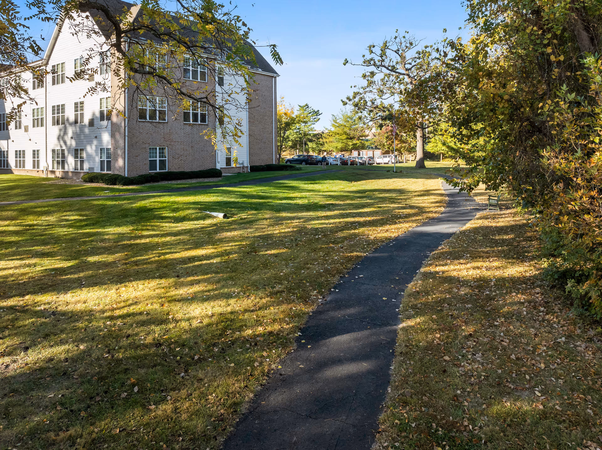 A paved walking path curves through a grassy area with scattered fallen leaves, leading towards a three-story building with white siding and brick accents. Trees with autumn foliage surround the area, and parked cars are visible in the background under a clear blue sky.