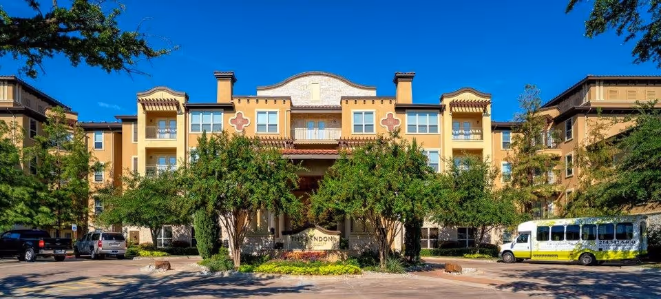 Front exterior view of The Landon at Lake Highlands senior living facility with a multi-story building, trees, parked cars, and a shuttle bus in front under a clear blue sky.