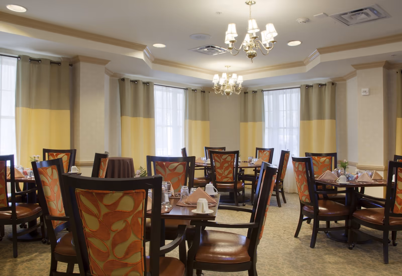 A dining room with multiple round wooden tables set with napkins, cups, glasses, and silverware. The chairs have dark wooden frames with orange and green patterned upholstery. The room has large windows with beige and yellow curtains, beige walls, and two chandeliers hanging from the ceiling.