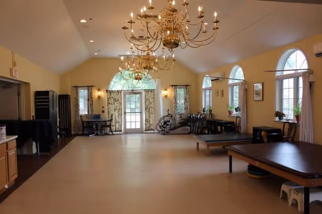 A spacious room with high ceilings featuring two large ornate chandeliers. The room has several arched windows with curtains, allowing natural light to enter. There are tables and chairs along the walls, exercise equipment near the windows, and a door with glass panels at the far end. The walls are painted a soft yellow, and the floor is a light beige color.
