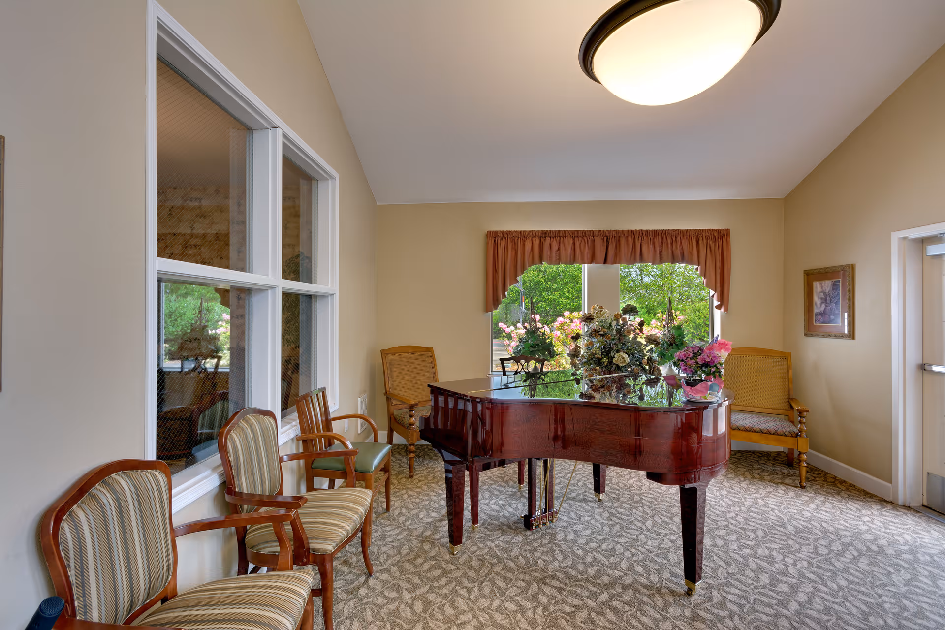 A bright room with a polished grand piano in the center, decorated with flower arrangements on top. The room has beige walls, a patterned carpet, and several wooden chairs with cushions arranged along the walls. A large window with a red valance shows greenery outside, and a round ceiling light illuminates the space.