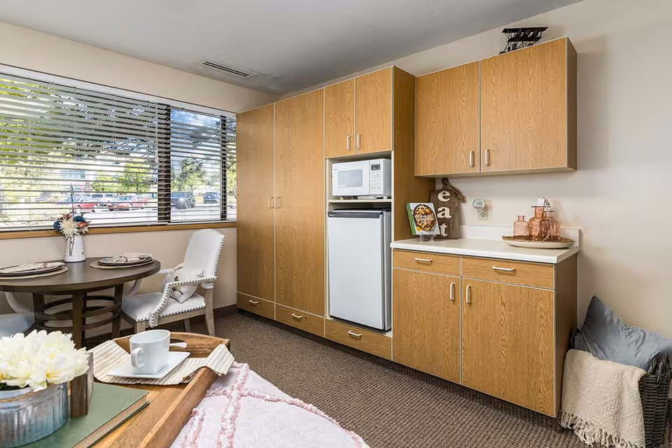 A small kitchen area in an assisted living facility with wooden cabinets, a microwave, and a mini refrigerator. Next to the kitchen is a round dining table set with plates and napkins, surrounded by upholstered chairs. A large window with blinds lets in natural light, and a cozy seating area with a tray holding a cup and flowers is partially visible in the foreground.