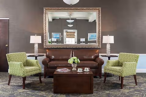 Lobby-style living room with brown leather sofa, green chairs, lamps, and a flower centerpiece.