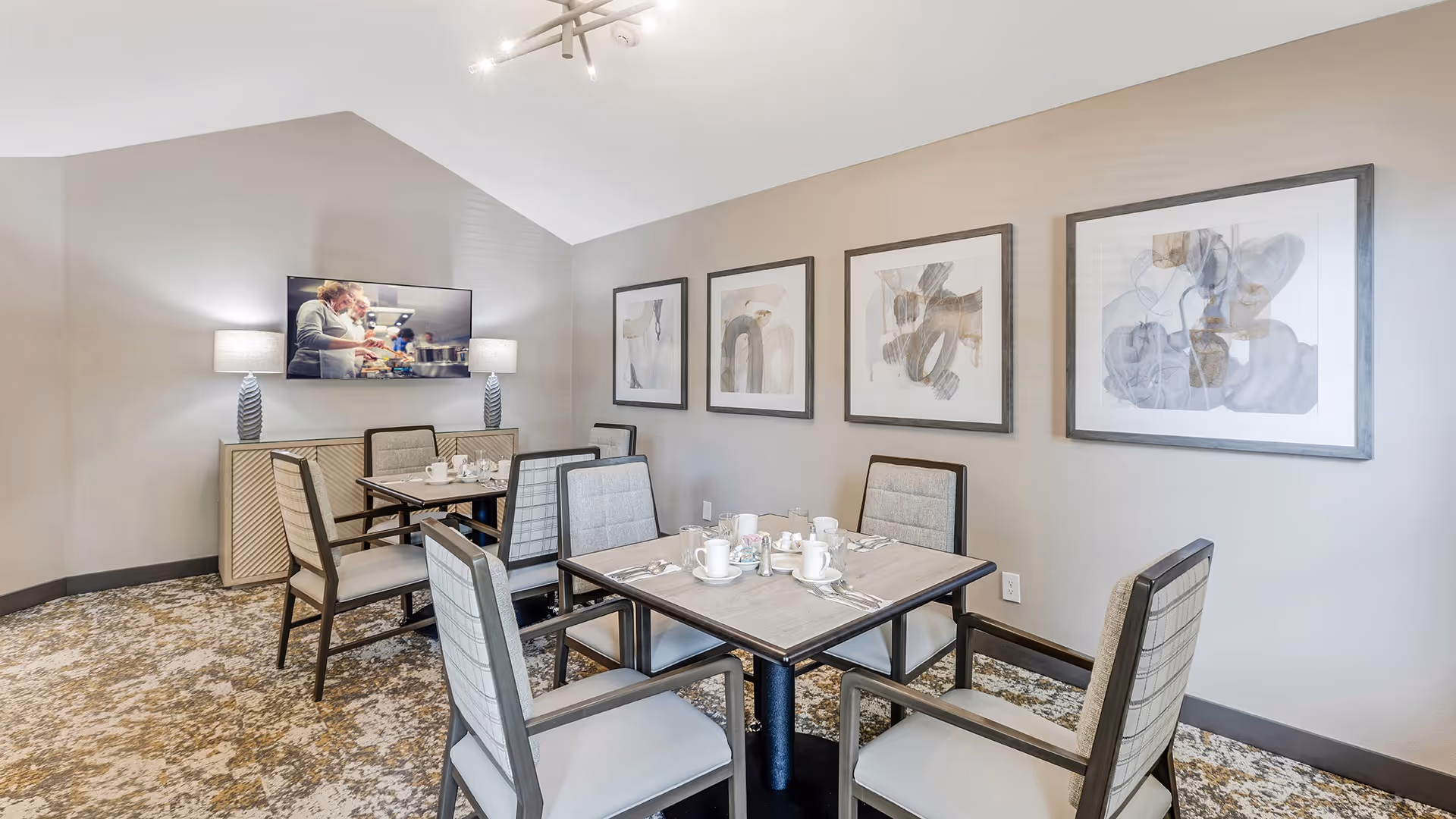 A dining area with two square tables, each set with white cups, saucers, and silverware. The room has six cushioned chairs with armrests around the tables. On the wall, there are four abstract art paintings in black frames. A flat-screen TV is mounted on the opposite wall above a sideboard with two table lamps. The carpet has a patterned design, and the walls are painted a light beige color.