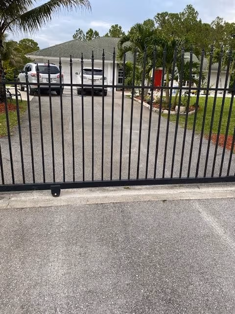 View of a gated driveway leading to a single-story residential building with a gray roof, two parked white vehicles, palm trees, and landscaped garden areas.