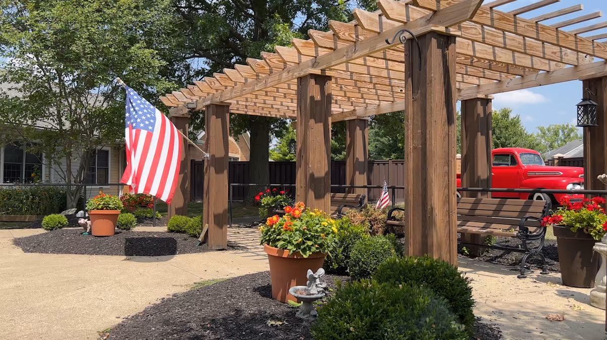 Outdoor courtyard with a wooden pergola, benches, potted flowers, an American flag, and a red vintage truck in the background.